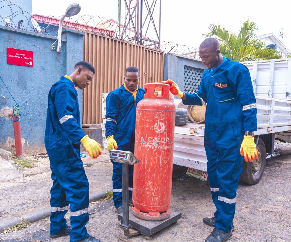 Three Suez Gas crew in branded coveralls and gloves weighing a cylinder on a digital scale beside their delivery truck.