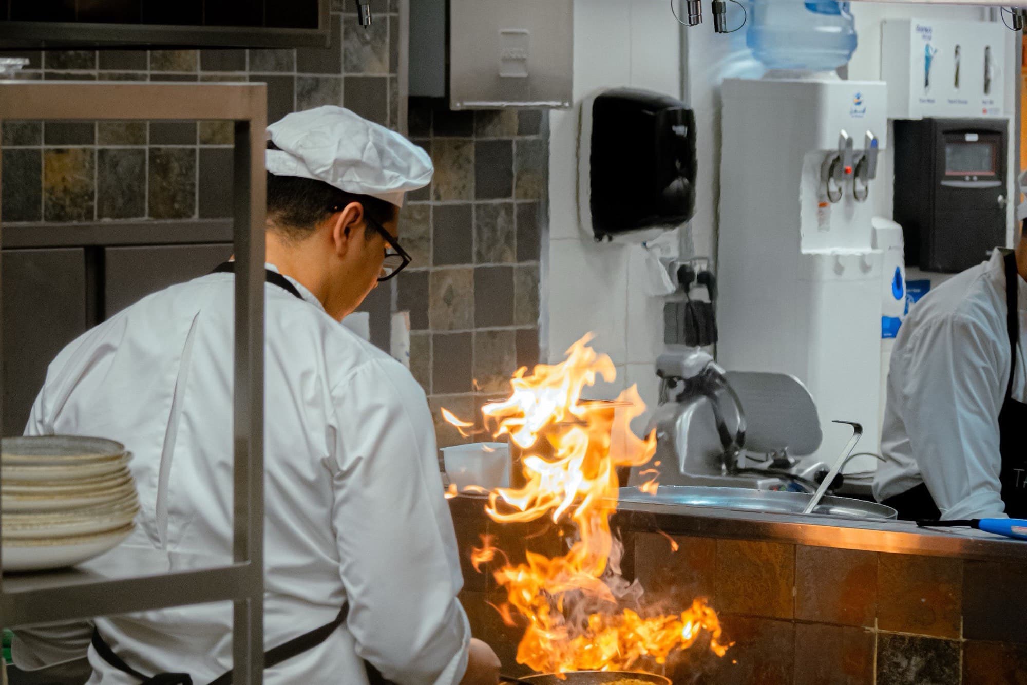 A chef working over a lit gas burner in a busy commercial kitchen, flame visible under the pan.